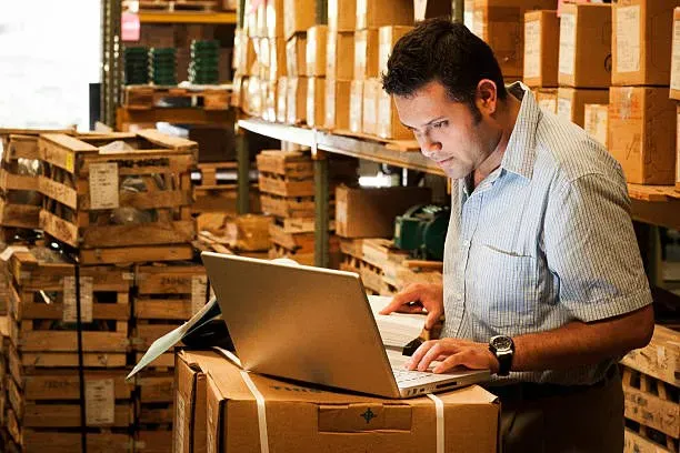 Warehouse worker using a laptop beside shelves of boxes